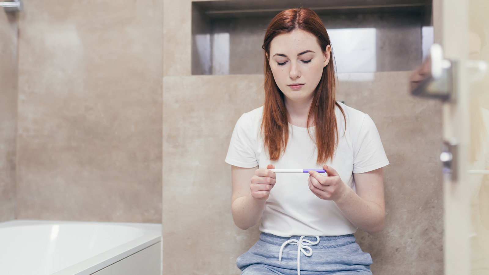 kobieta w wieku 30 plus siedzi w łazience i trzyma w ręku test ciążowy z poważną miną