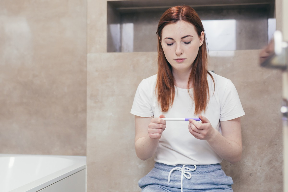 kobieta w wieku 30 plus siedzi w łazience i trzyma w ręku test ciążowy z poważną miną