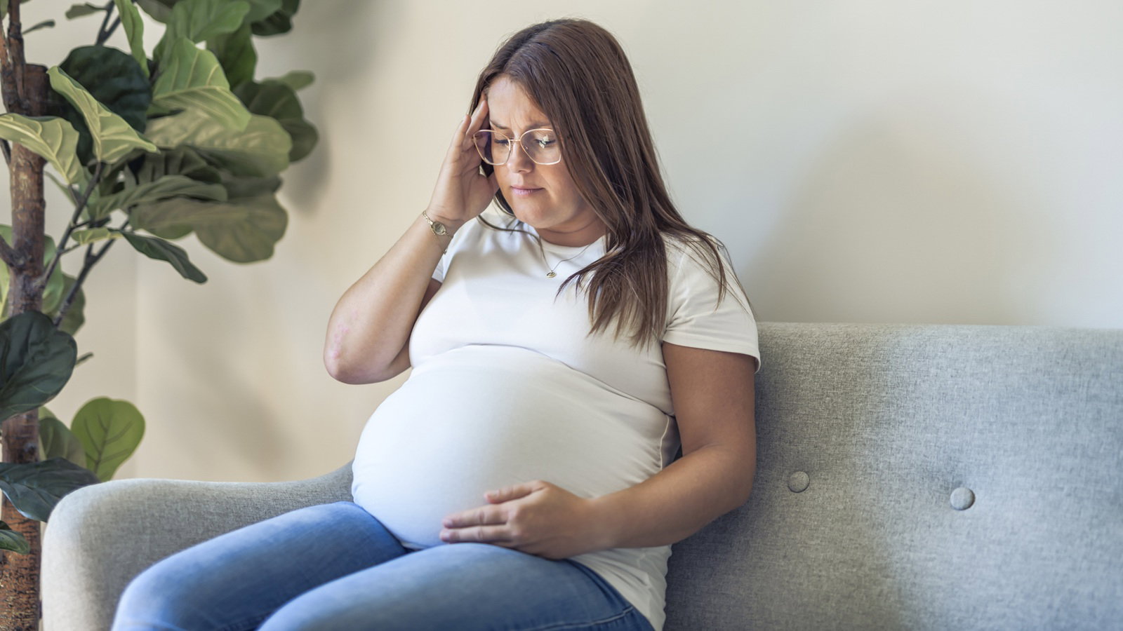 kobieta w wieku 30 plus w białym t-shircie z ciążowym brzuszkiem i w okularach siedzi zmartwiona i załamana na kanapie