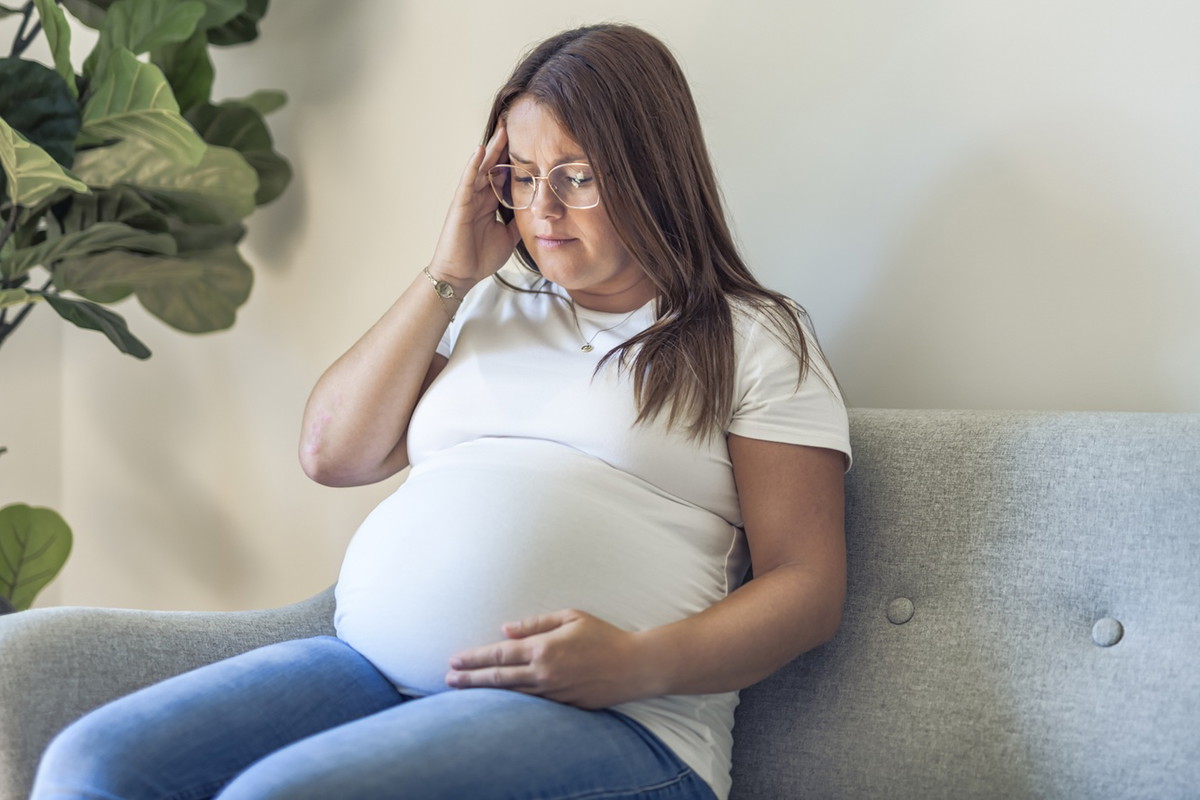kobieta w wieku 30 plus w białym t-shircie z ciążowym brzuszkiem i w okularach siedzi zmartwiona i załamana na kanapie