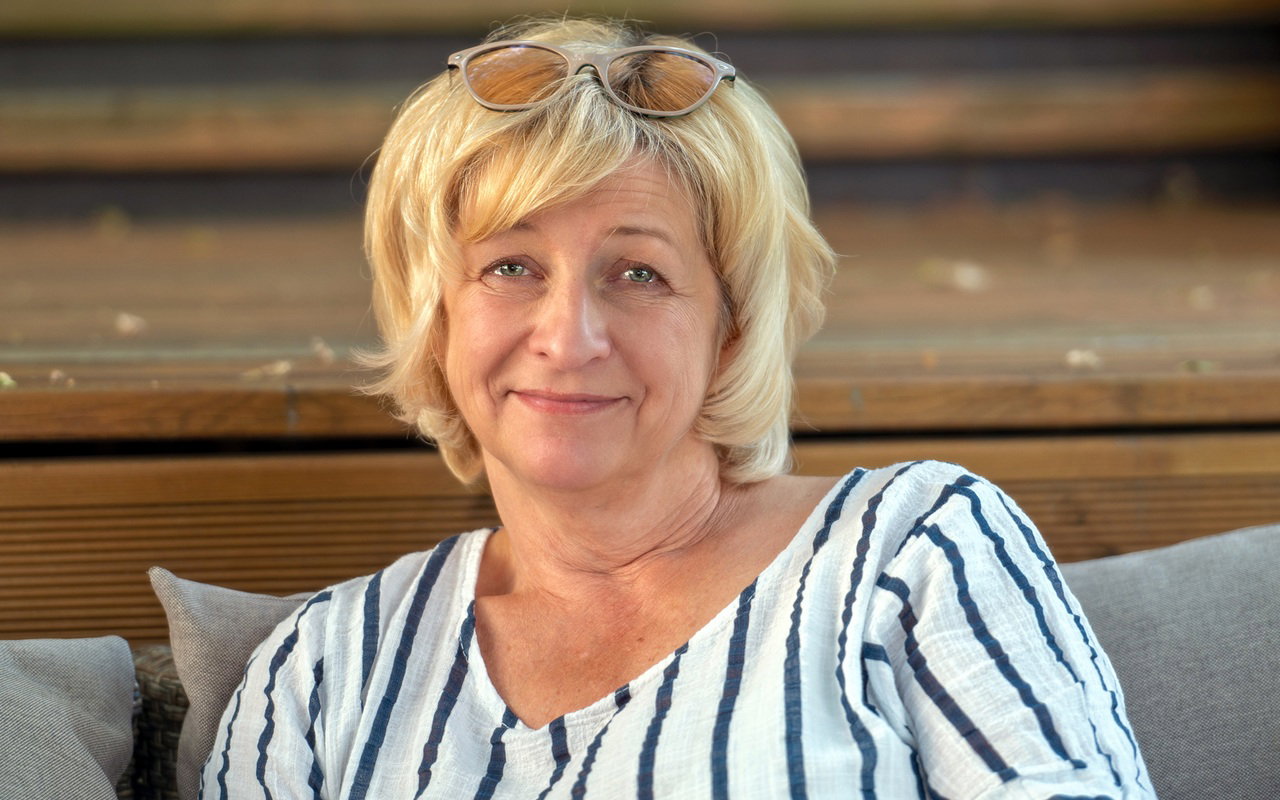 kobieta w wieku 55 plus blondynka  w pasiatej bluzce z kpiącym i lekko smutnym uśmieszkiem siedzi na tarasie