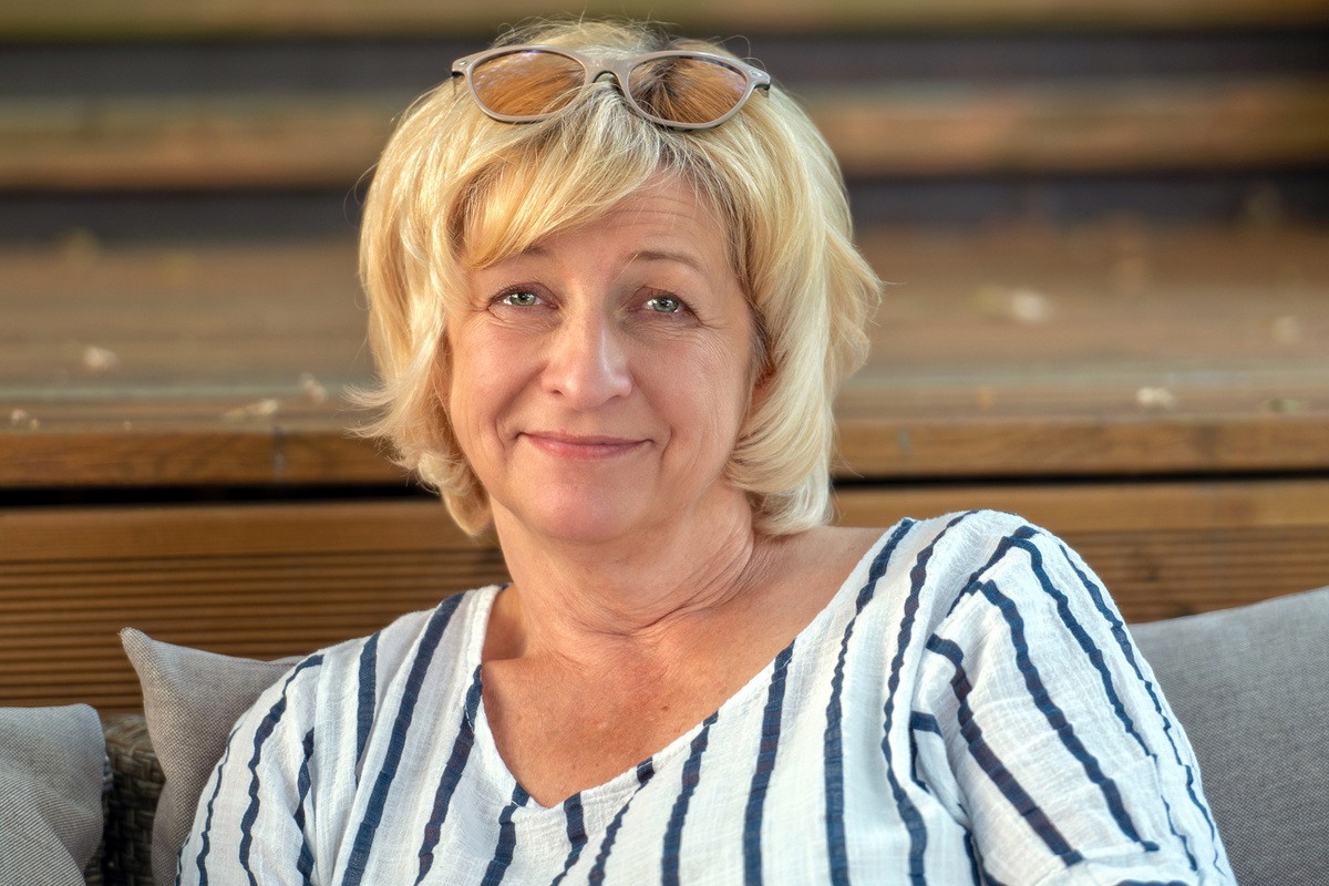 kobieta w wieku 55 plus blondynka w pasiatej bluzce z kpiącym i lekko smutnym uśmieszkiem siedzi na tarasie