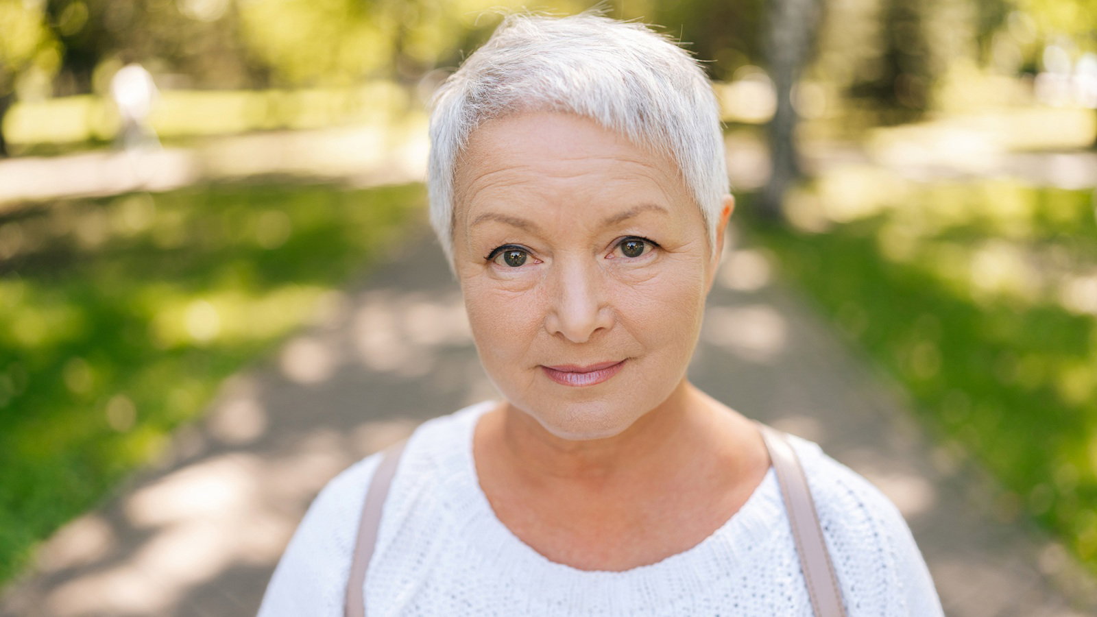 kobieta w wieku 65 plus z swiymi krótkimi włosami stoi na tle parku zamyślona z lekkim uśmiechem i nadzieją w oczach