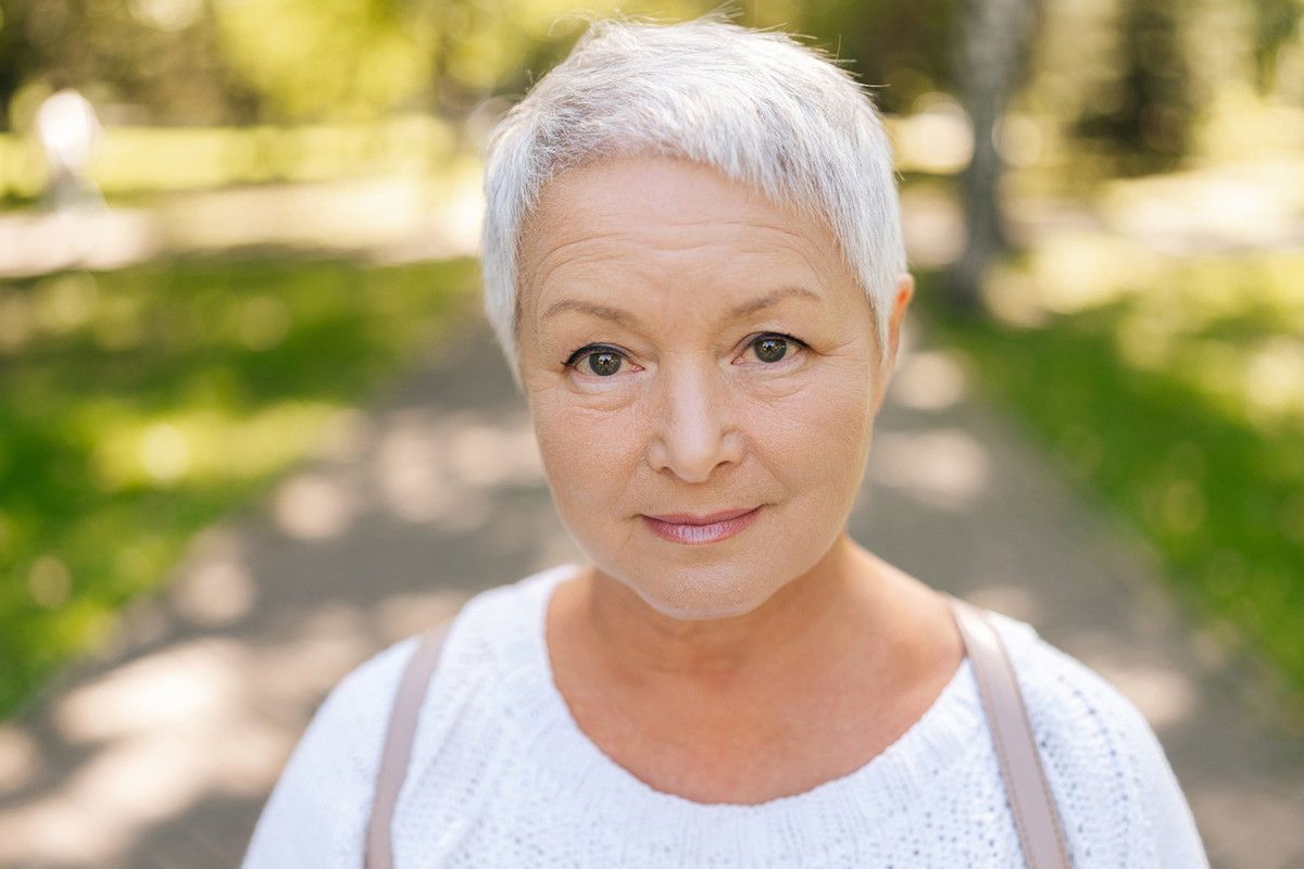 kobieta w wieku 65 plus z swiymi krótkimi włosami stoi na tle parku zamyślona z lekkim uśmiechem i nadzieją w oczach