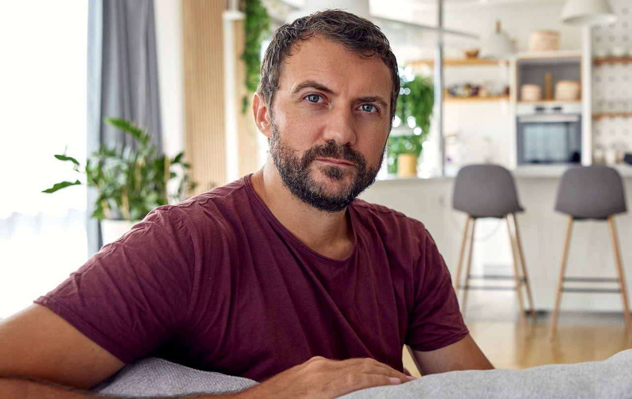 mężczyzna w wieku 30 plus z zarostem siedzi w domu na kanapie w brązowym t-shircie zamyślony