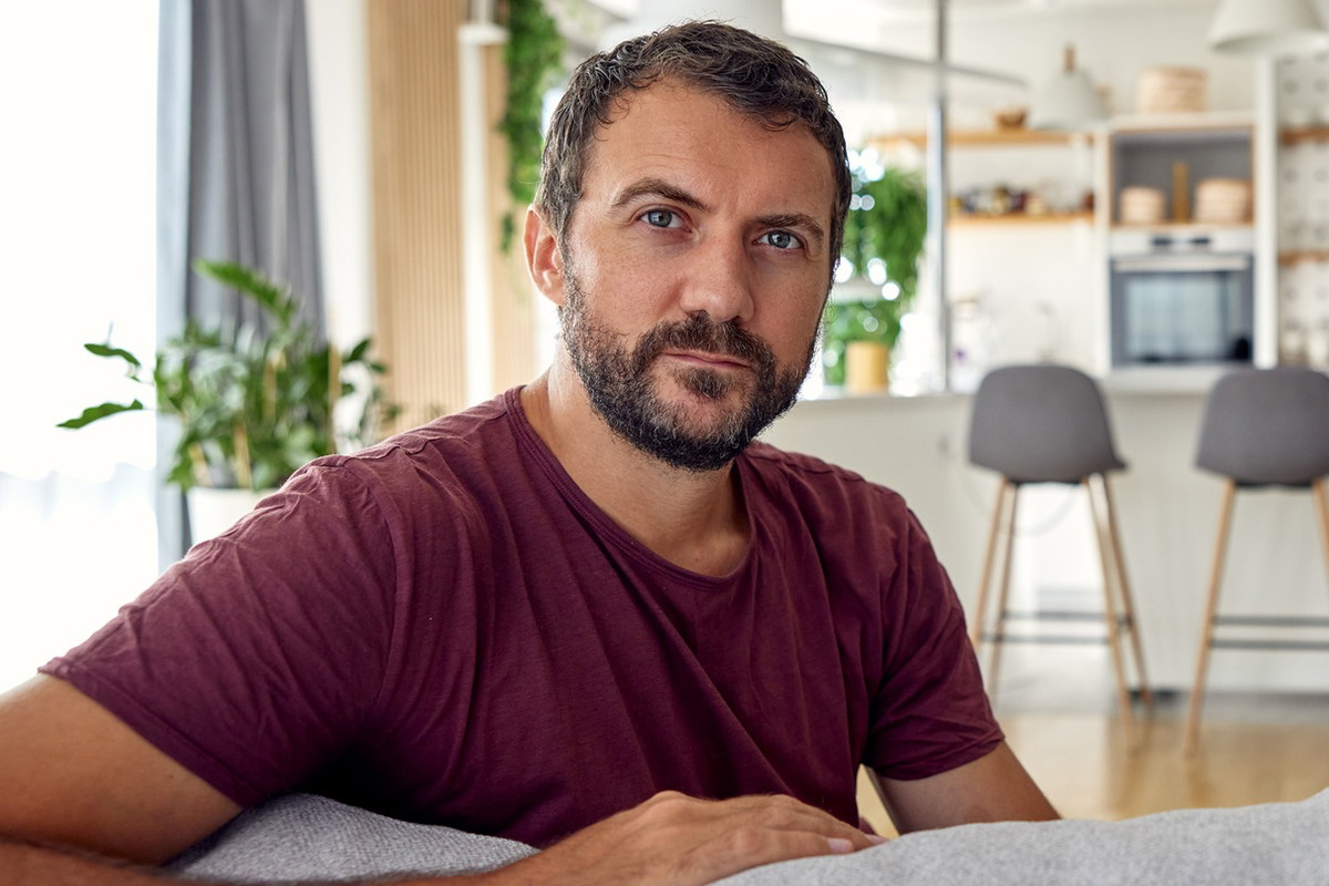 mężczyzna w wieku 30 plus z zarostem siedzi w domu na kanapie w brązowym t-shircie zamyślony