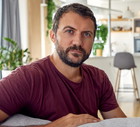 mężczyzna w wieku 30 plus z zarostem siedzi w domu na kanapie w brązowym t-shircie zamyślony