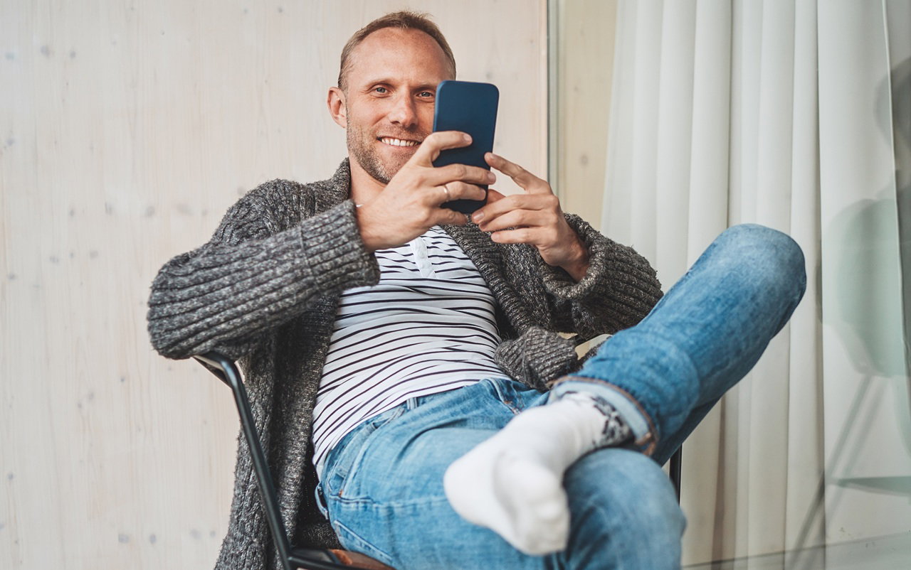 mężczyzna w wieku 35 plus w szarym swetrze siedzi z telefonem w ręku w hotelu i się uśmiecha coś oglądając