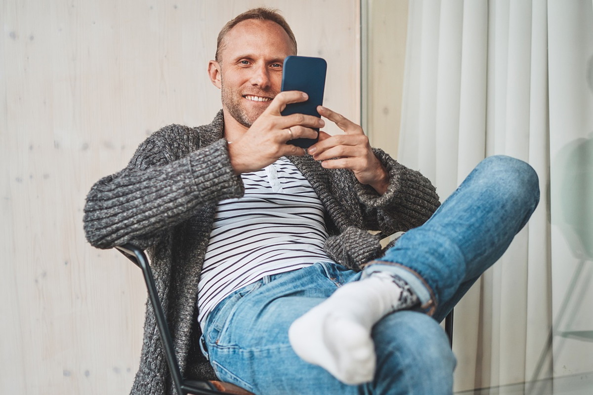 mężczyzna w wieku 35 plus w szarym swetrze siedzi z telefonem w ręku w hotelu i się uśmiecha coś oglądając