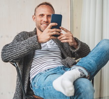 mężczyzna w wieku 35 plus w szarym swetrze siedzi z telefonem w ręku w hotelu i się uśmiecha coś oglądając