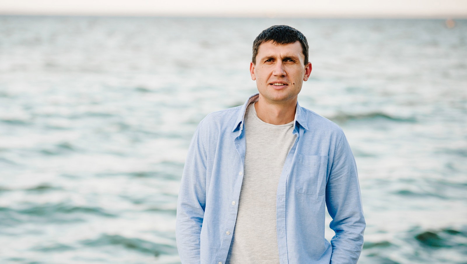 mężczyzna w wieku 40 plus stoi na plaży w niebieskiej koszuli z poważną miną