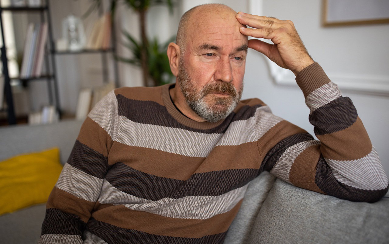 meżczyzna w wieku 50 plus łysiejący siedzi na kanapie w swetrze w pasy zamyślony i załamany