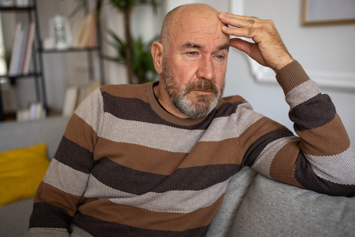 meżczyzna w wieku 50 plus łysiejący siedzi na kanapie w swetrze w pasy zamyślony i załamany