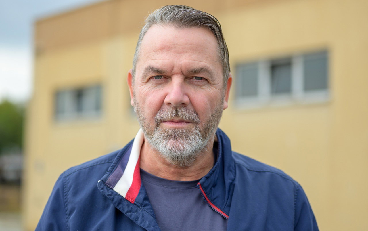 mężczyzna w wieku 55 plus siwiejący z brodą w granatowej kurtce stoi na tle żółtego budynku z poważną miną