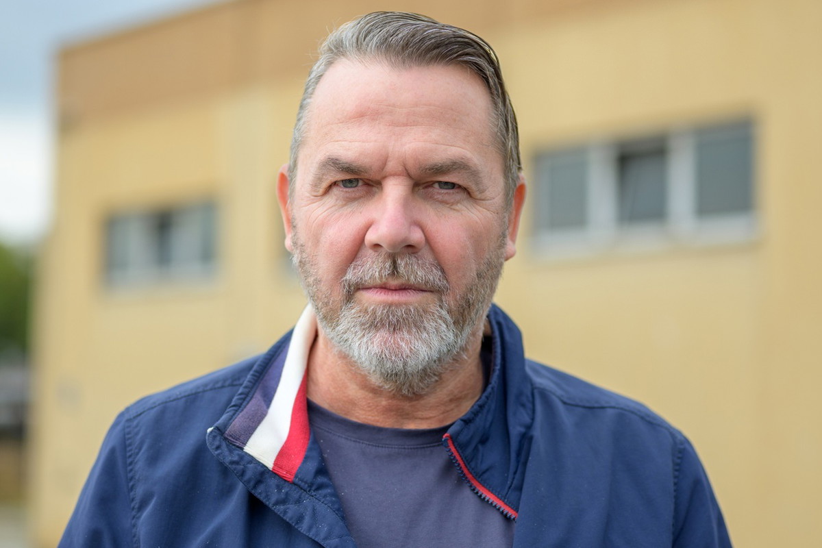 mężczyzna w wieku 55 plus siwiejący z brodą w granatowej kurtce stoi na tle żółtego budynku z poważną miną