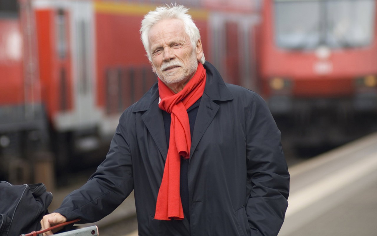 mężczyzna w wieku 65 plus siwy z wąsami i zarostem w czerwonym szaliku stoi na stacji kolejowej z poważną miną