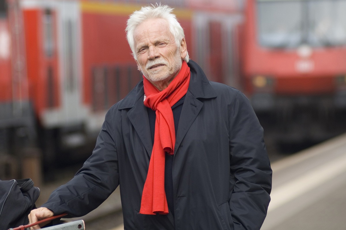 mężczyzna w wieku 65 plus siwy z wąsami i zarostem w czerwonym szaliku stoi na stacji kolejowej z poważną miną
