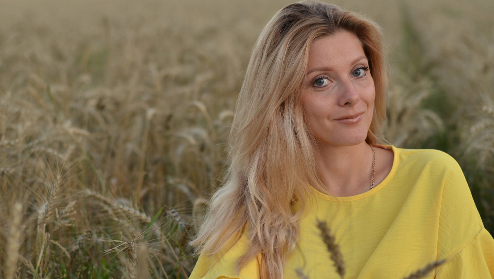 kobieta w wieku 25 plus blondynka w żółtej bluzce siedzi na tle zboża z lekkim uśmiechem
