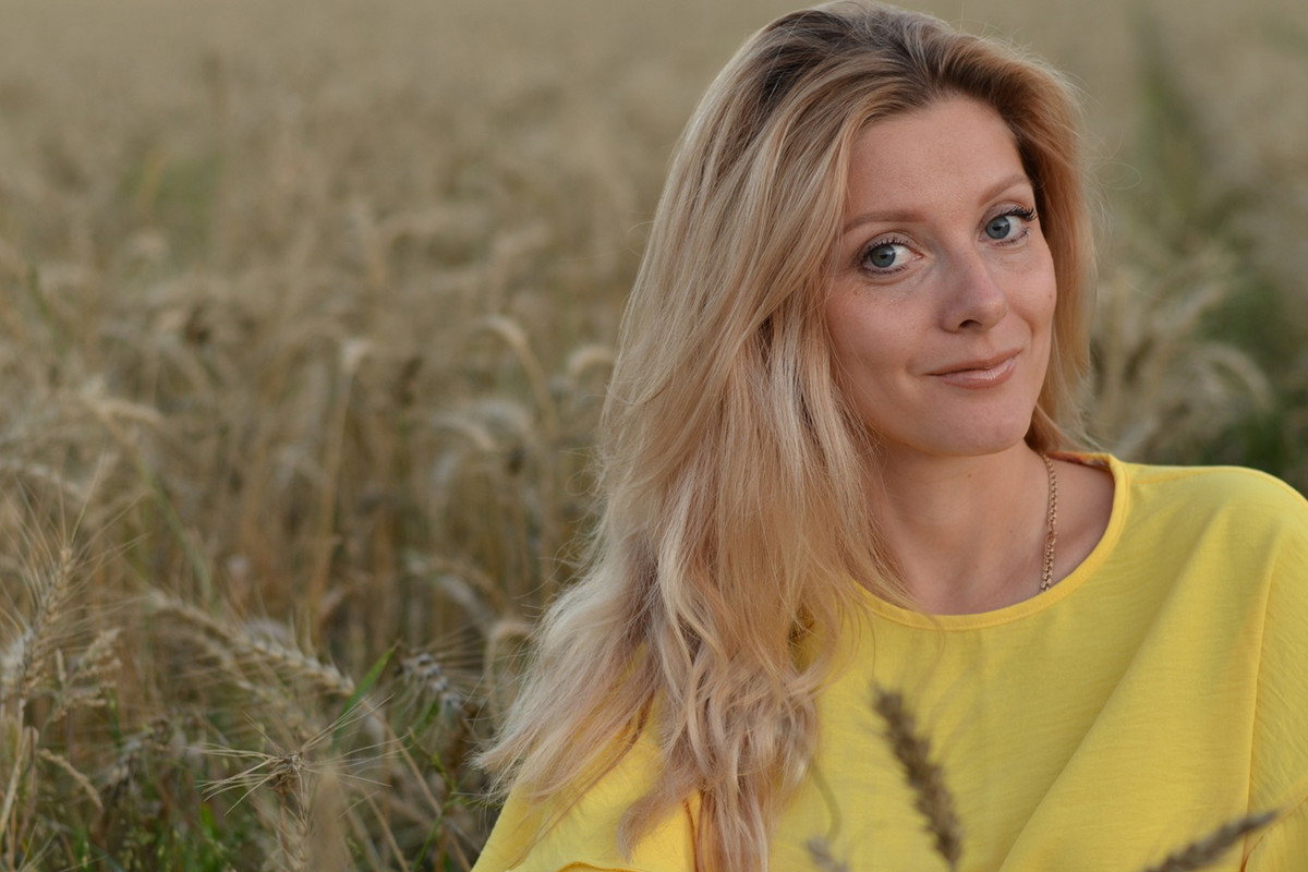 kobieta w wieku 25 plus blondynka w żółtej bluzce siedzi na tle zboża z lekkim uśmiechem