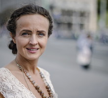kobieta w wieku 35 plus w białej koronkowej sukience i w koralach stoi na ulicy z poważną miną
