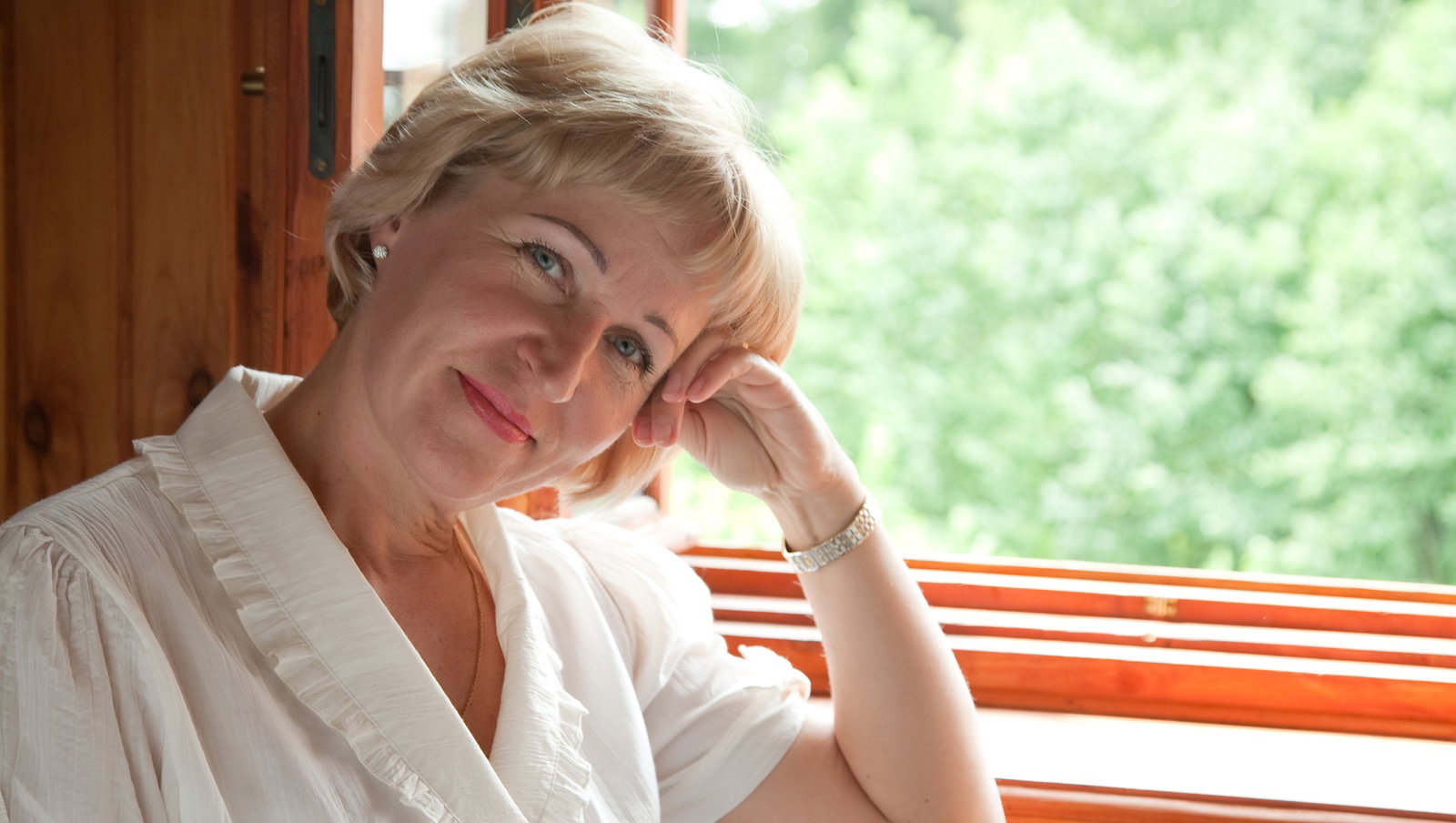 kobieta w wieku 45 plus blondynka siedzi przy oknie w białym stroju rozmarzona i zamyślona