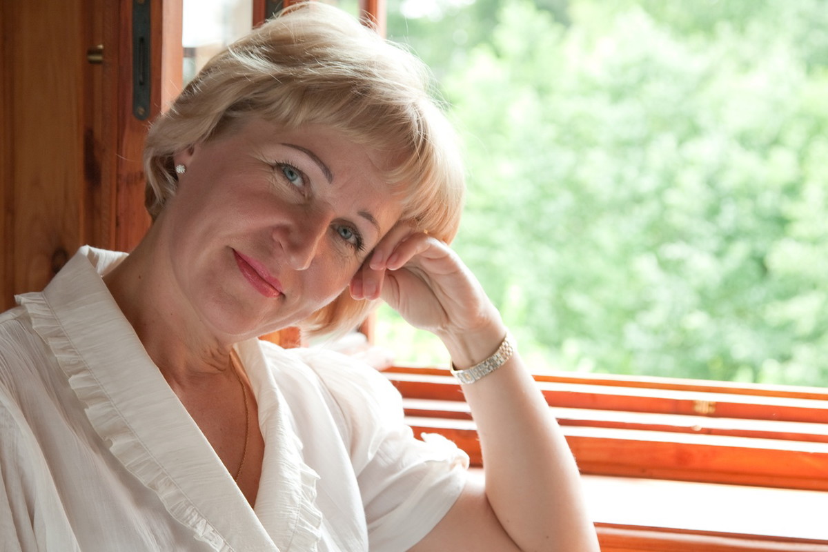 kobieta w wieku 45 plus blondynka siedzi przy oknie w białym stroju rozmarzona i zamyślona