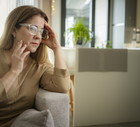 kobieta w wieku 50 plus w okularach siedzi na tle sprzątniętego mieszkania zamyślona i smutna