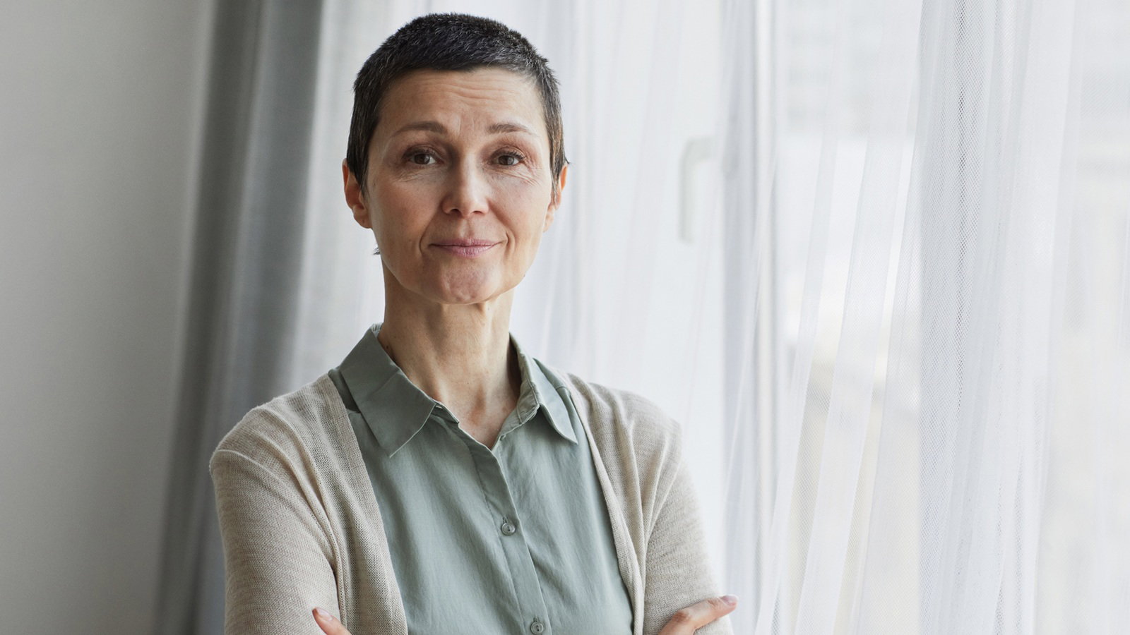 kobieta w wieku 50 plus z krótkimi włosami stoi przy oknie ze smutnym uśmiechem zamyślona