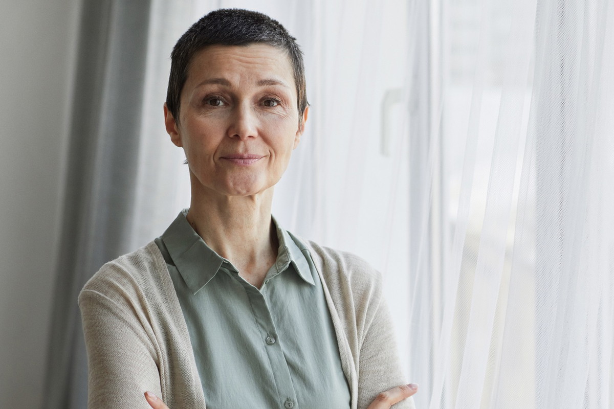 kobieta w wieku 50 plus z krótkimi włosami stoi przy oknie ze smutnym uśmiechem zamyślona