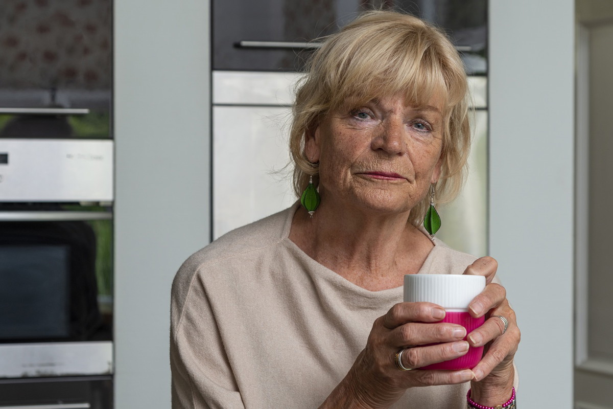 kobieta w wieku 65 plus blondynka siedzi w kuchni z kubkiem w ręce zamyślona i smutna