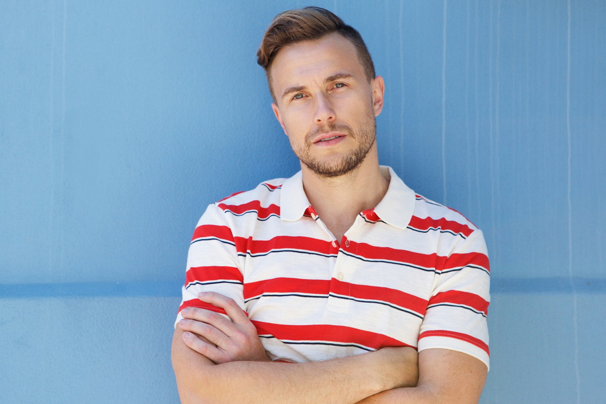 mężczyzna w wieku 30 plus blondyn w koszulce w pasy na tle niebieskiej ściany stoi zamyślony