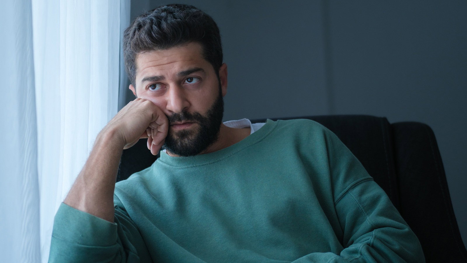 mężczyzna w wieku 30 plus z brodą i wąsami w zielonym swetrze siedzi przy oknie zamyślony i smutny