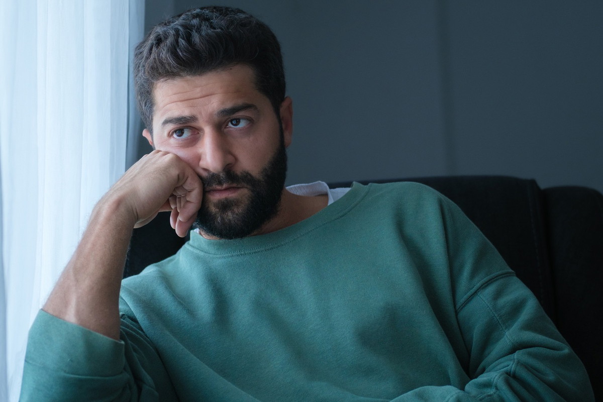 mężczyzna w wieku 30 plus z brodą i wąsami w zielonym swetrze siedzi przy oknie zamyślony i smutny