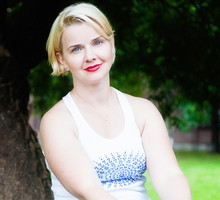 kobieta w wieku 30 plus blondynka w białej koszulce siedzi na tle zieleni z lekkim uśmiechem
