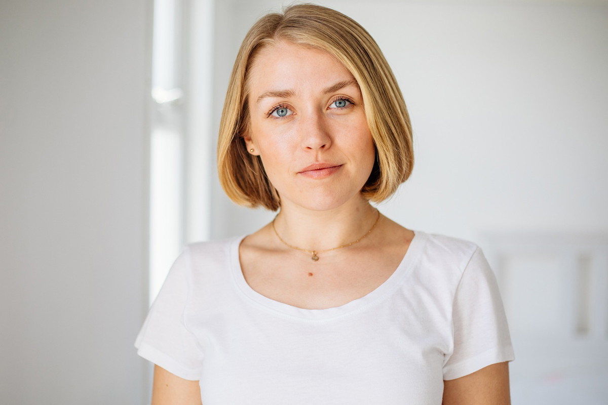 kobieta w wieku 30 plus blondynka w białym t-shircie z lekkim uśmiechem stoi an tle jasnego pomieszczenia