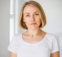 kobieta w wieku 30 plus blondynka w białym t-shircie z lekkim uśmiechem stoi an tle jasnego pomieszczenia