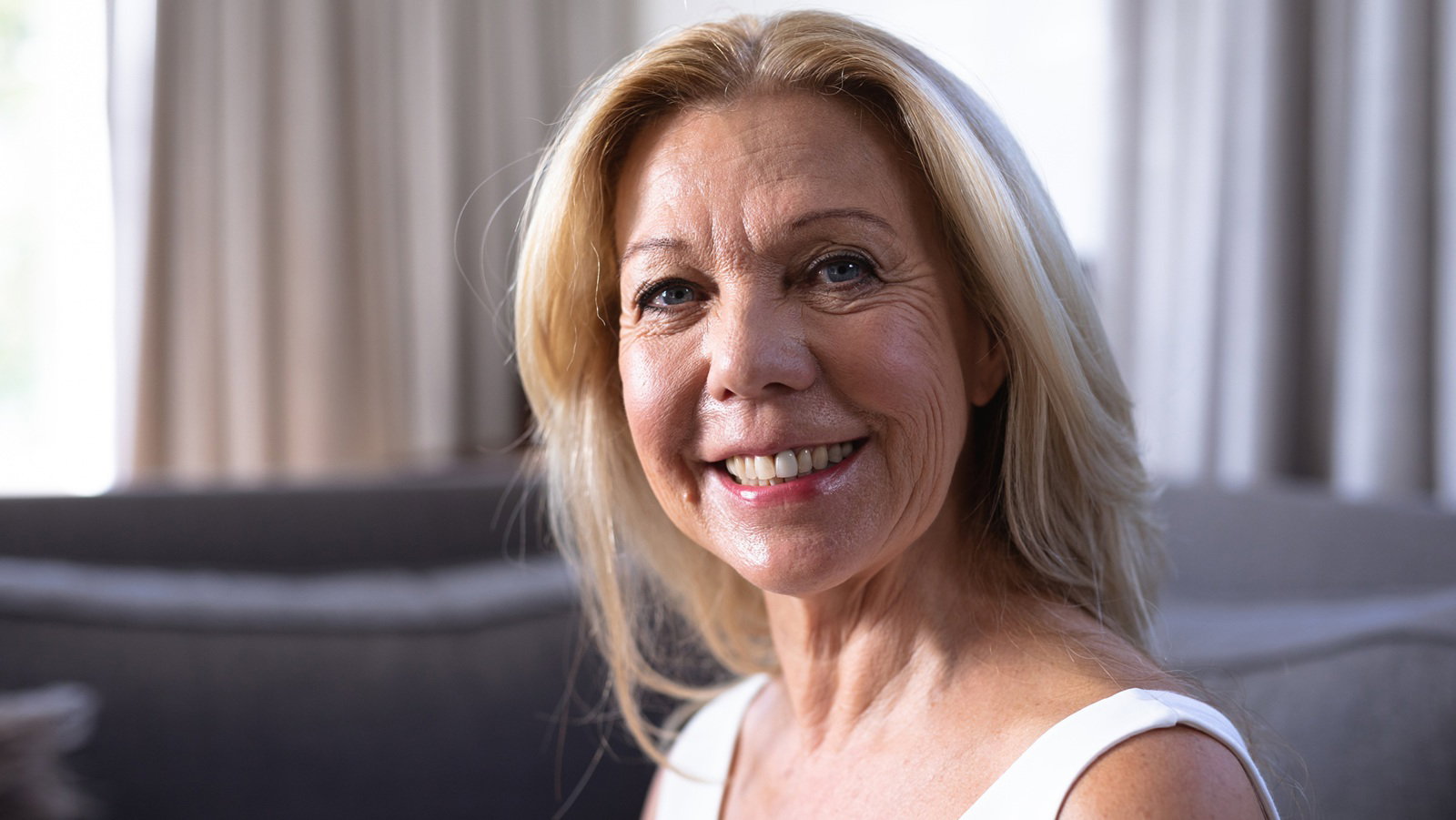 kobieta w wieku 60 plus blondynka siedzi na kanapie na tle okna i szeroko się uśmiecha