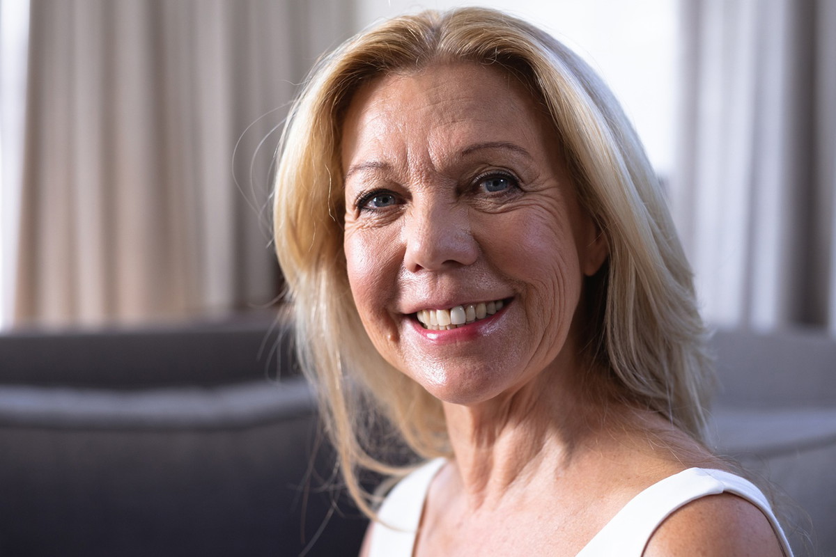kobieta w wieku 60 plus blondynka siedzi na kanapie na tle okna i szeroko się uśmiecha