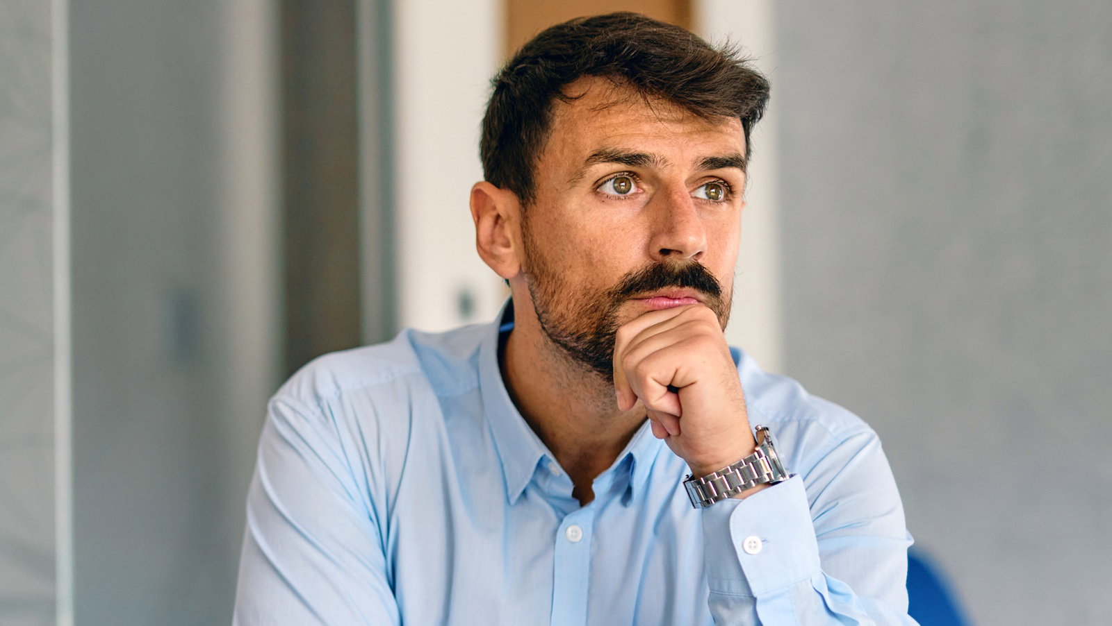 mężczyzna w wieku 35 plus z ciemnym zarostem i w błękitnej koszuli siedzi przy stole zamyślony