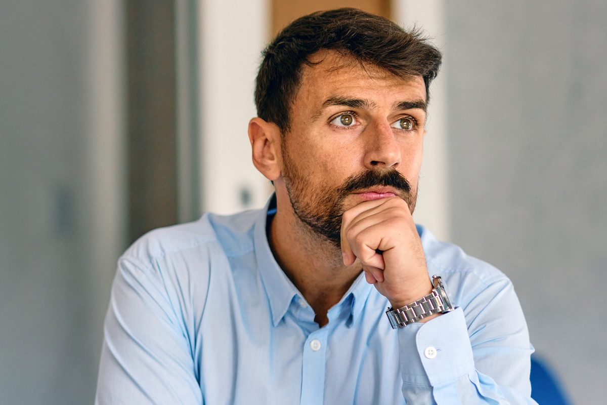mężczyzna w wieku 35 plus z ciemnym zarostem i w błękitnej koszuli siedzi przy stole zamyślony