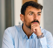 mężczyzna w wieku 35 plus z ciemnym zarostem i w błękitnej koszuli siedzi przy stole zamyślony