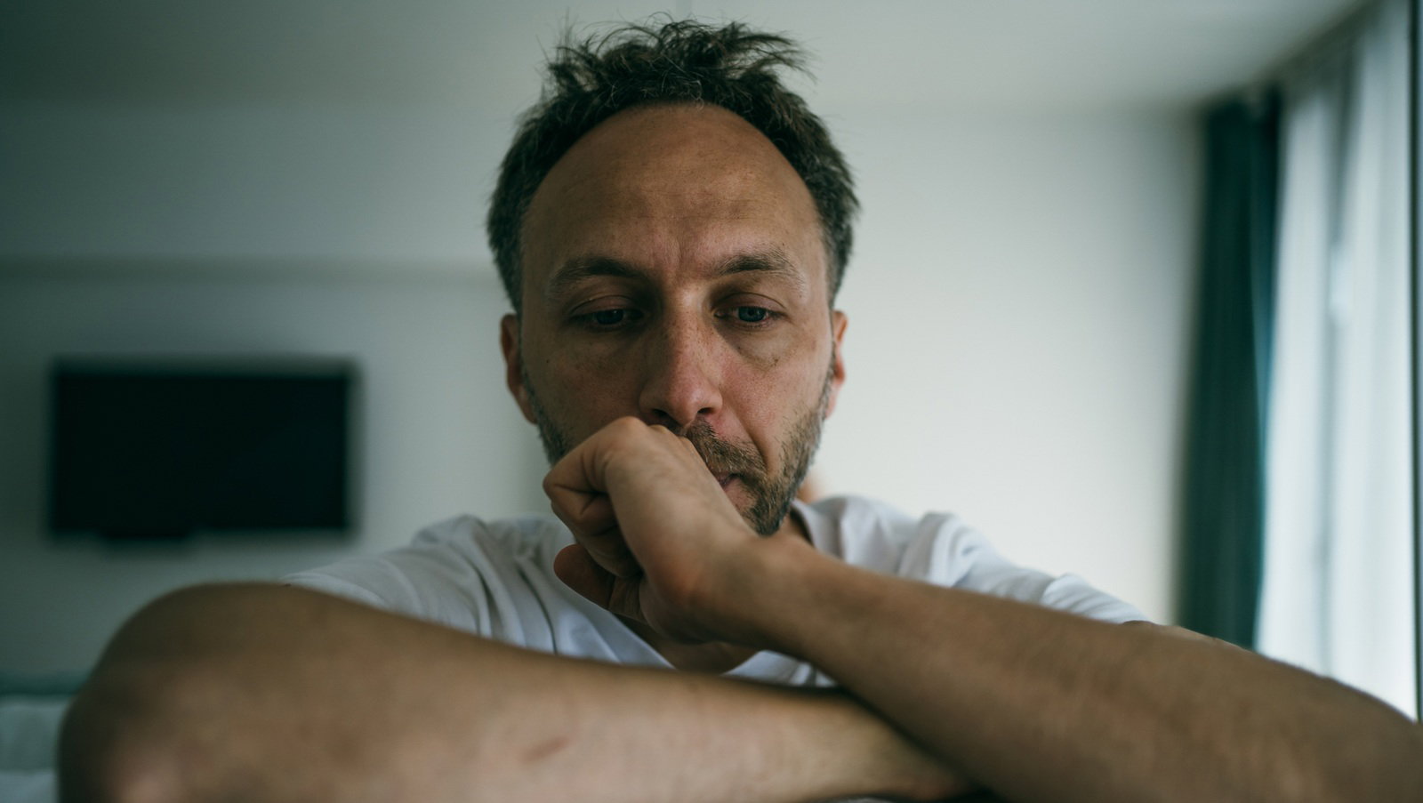 mężczyzna w wieku 35 plus z zarostem łysiejący w białej koszulce siedzi załamany i smutny na tle salonu