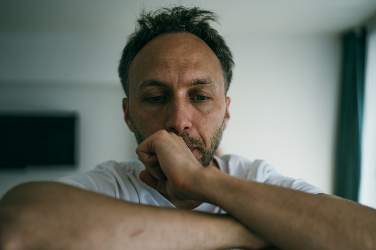 mężczyzna w wieku 35 plus z zarostem łysiejący w białej koszulce siedzi załamany i smutny na tle salonu