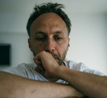 mężczyzna w wieku 35 plus z zarostem łysiejący w białej koszulce siedzi załamany i smutny na tle salonu