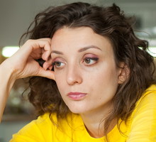 kobieta w wieku 30 plus w żółtej koszulce i kręconymi włosami siedzi przy stole w kuchni zamyślona i smutna