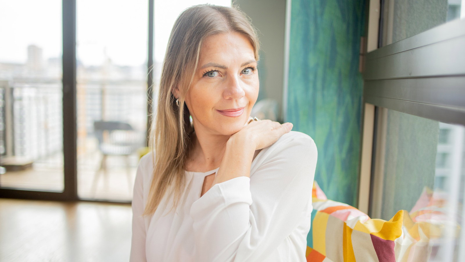kobieta w wieku 35 plus blondynka w białej sukience siedzi na tle dużych okien z zawadiackim uśmiechem