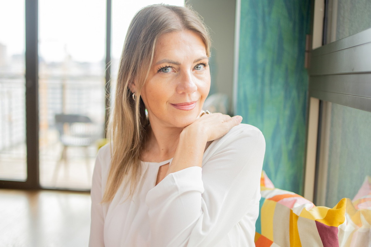kobieta w wieku 35 plus blondynka w białej sukience siedzi na tle dużych okien z zawadiackim uśmiechem