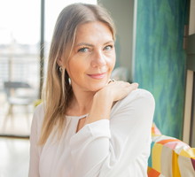 kobieta w wieku 35 plus blondynka w białej sukience siedzi na tle dużych okien z zawadiackim uśmiechem