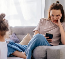 kobieta w wieku 35 plus siedzi na kanapie z telefonem w ręku i patrzy na swoje dziecko bez uśmiechu