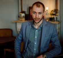 mężczyzna w wieku 30 plus z zarostem łysiejący stoi na tle mebli w stylu retro w marynarce z poważną miną